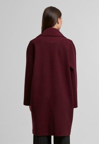 Abrigo de mezcla de lana burdeos con un amplio cuello, corte recto, mangas largas y bolsillos laterales. Textura suave con un diseño minimalista.
