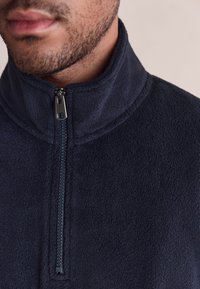 Marineblauer Fleece-Pullover mit hohem Kragen und halbem Reißverschluss; verfügt über eine weiche Textur und sichtbare Nähte entlang der Kanten.