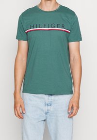 Camiseta de algodón verde con cuello redondo, que presenta el texto "HILFIGER" y un acento a rayas rojas y blancas en el pecho. Manga corta.