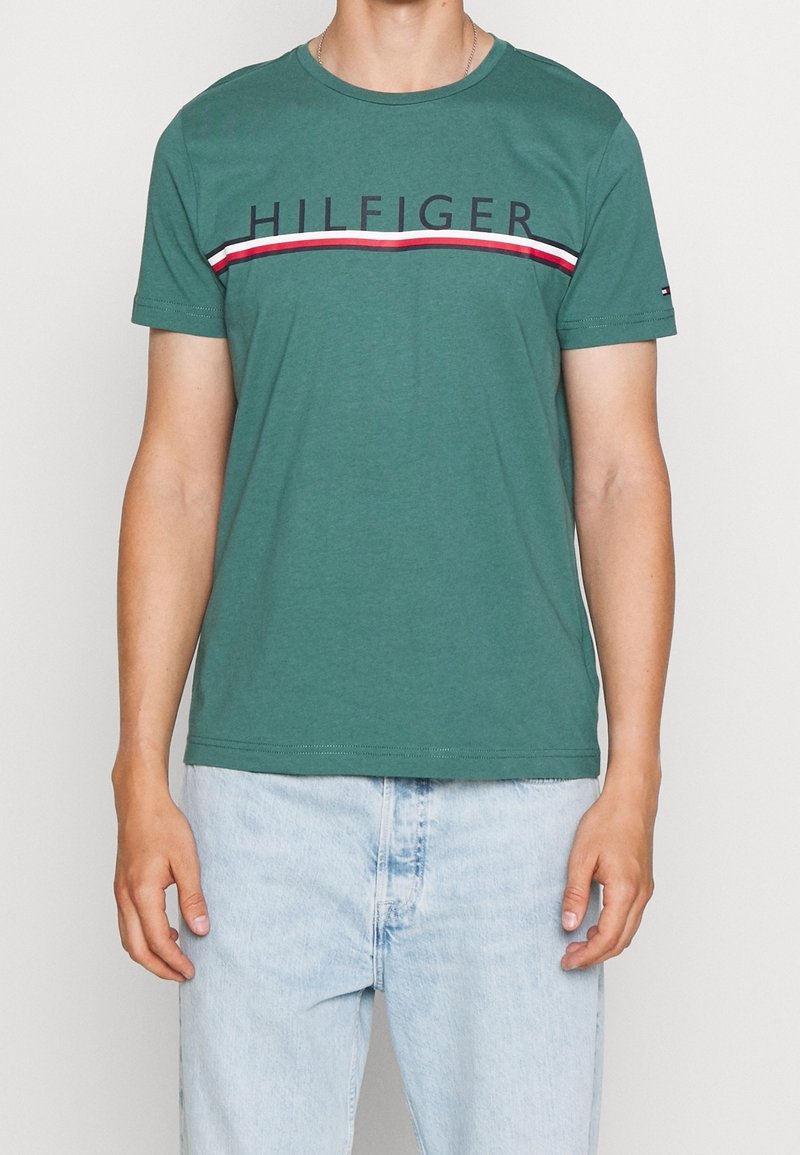 Camiseta de algodón verde con cuello redondo, que presenta el texto "HILFIGER" y un acento a rayas rojas y blancas en el pecho. Manga corta.