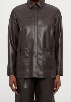 Ciemnobrązowa kurtka z ekoskóry z kieszeniami na przód zapinanymi na zatrzaski i szpiczasty kołnierz, noszona z dopasowanymi spodniami.