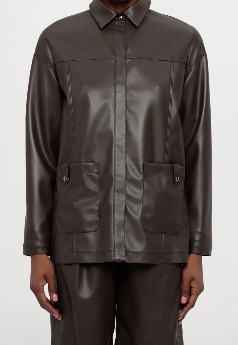 Veste en similicuir marron foncé avec poches avant à boutons-pression et col pointu, portée avec un pantalon assorti.