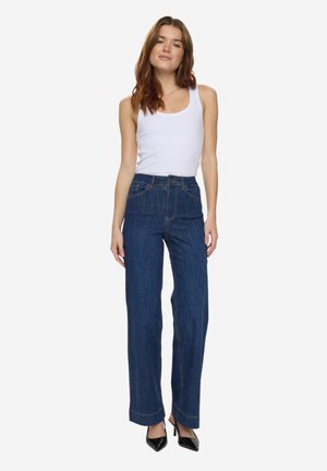 Femeie cu păr lung și castaniu, purtând un top alb fără mâneci, blugi largi cu talie înaltă, de culoare albastru închis, și pantofi negri cu vârf ascuțit.