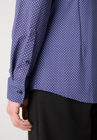 Camisa de patrones azul y blanco con pequeños cuadrados, con puños abotonados y cola recta, llevada sobre pantalones de tela negra.