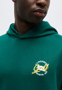 Artimas asmuo, dėvintis žalią džemperį su gaubtu, ant krūtinės esantis geltonos ir baltos spalvos logotipas, kuriame užrašyta „J. Lindeberg Golf Augusta, GA“.