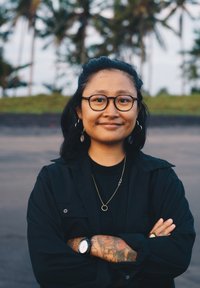 Inimene, kes kannab musta nööpidega särki musta t-särgi peal, kuldset kaelakeed ja kella, käte peal on tätoveeringud, naeratab välistingimustes.