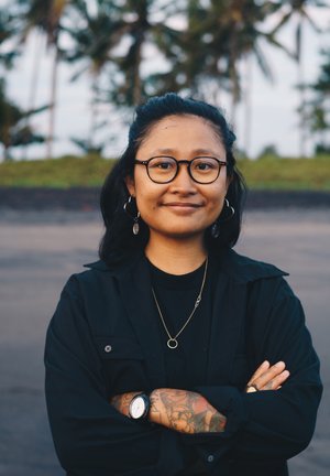 Inimene, kes kannab musta nööpidega särki musta t-särgi peal, kuldset kaelakeed ja kella, käte peal on tätoveeringud, naeratab välistingimustes.