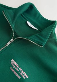 Grüner Reißverschluss-Sweatshirt mit einem gerippten Kragen, mit pinkem Text "STEREO IMAGING Waveform Studios AMSTERDAM." Glatte Stoffoberfläche.