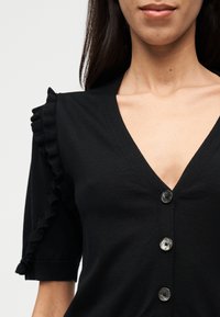 Μαύρο πλεκτό ζακέτα με V-λαιμόκοψη, κλείσιμο με κουμπιά και διακοσμητικές βολάν στα κοντά μανίκια. Λείο ύφασμα με διακριτική γυαλάδα.