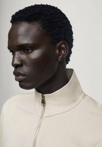 Sweatshirt crem cu fermoar, cu guler înalt, dintr-un material texturat, și un fermoar argintiu care subliniază designul curat. Pielea închisă la culoare a modelului contrastează puternic.