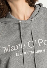 Sudadera gris con capucha que presenta un cordón con herrajes plateados, impresa con "Marc O'Polo EST. N STOCKHOLM" en texto blanco en la parte frontal.