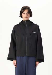 Veste noire à capuche avec fermeture éclair à l'avant, deux poches poitrine et poignets en Velcro. Présente une coupe décontractée et un design minimaliste avec un logo de marque.