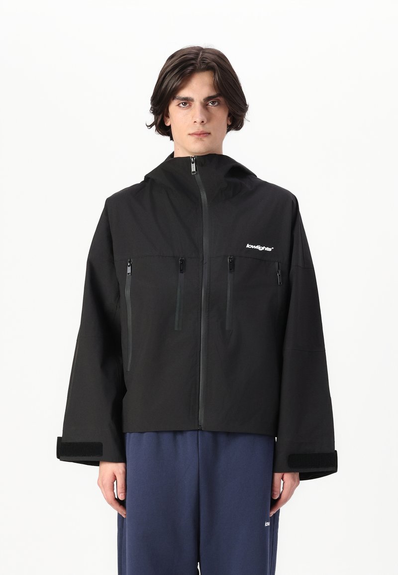 Veste noire à capuche avec fermeture éclair à l'avant, deux poches poitrine et poignets en Velcro. Présente une coupe décontractée et un design minimaliste avec un logo de marque.