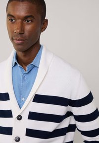 Cardigan bianco con strisce orizzontali blu navy, dotato di un colletto a scialle, tre bottoni e un design a maglia testurizzata. Indossato sopra una camicia azzurro chiaro.
