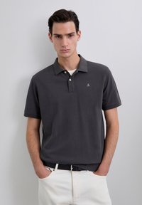 Polo en gris oscuro con cuello, que presenta un pequeño logo en el pecho. Combinado con pantalones beige claros y un cinturón negro.