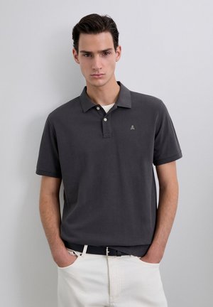 Polo en gris oscuro con cuello, que presenta un pequeño logo en el pecho. Combinado con pantalones beige claros y un cinturón negro.