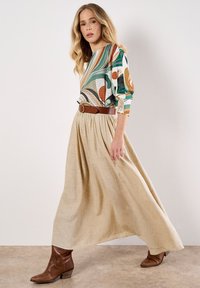 Una donna che cammina indossando un top a fantasia colorato, una maxi gonna beige con una cintura marrone e stivaletti con tacco marroni su uno sfondo neutro.