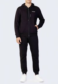 Moški v črni zip-up kapuci in ujemajočih se črnih trenirkah, obut v bele superge, stoji z eno roko v žepu.