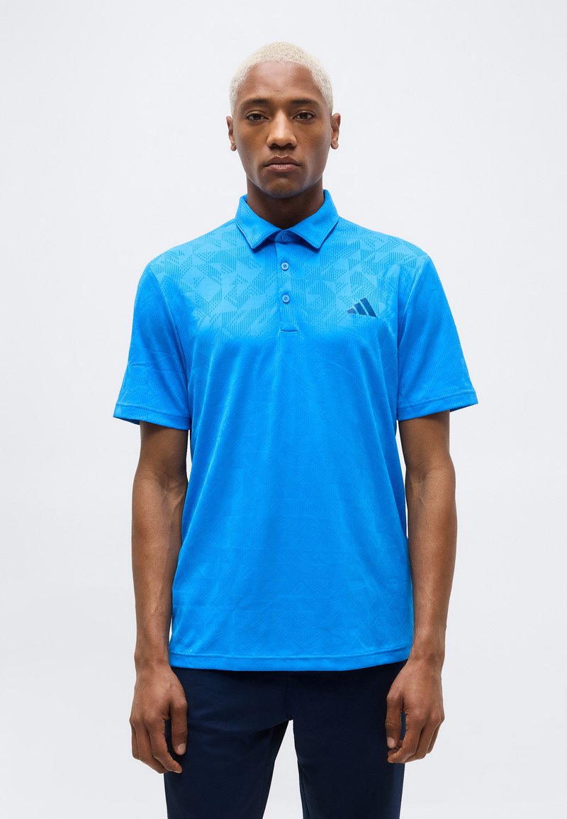 Νέος άνδρας με κοντά ξανθά μαλλιά που φοράει μπλε πουκάμισο πόλο με φωτεινά σχέδια και σκούρο παντελόνι, στέκεται μπροστά σε απλό λευκό φόντο.