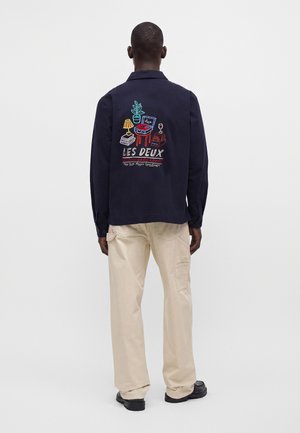 Mand iført navy jakke med farverigt broderet hjemmemotiv og beige bukser, stående med ryggen til mod en ensfarvet baggrund.