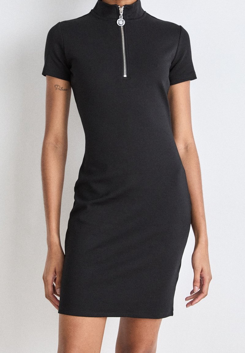 Czarna sukienka z krótkim rękawem, wykonana z elastycznego materiału, z wysokim kołnierzem i srebrnym zamkiem błyskawicznym z okrągłym uchwytem. Dopasowany krój.