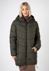 Frau in einer olivgrünen, gesteppten Winterjacke mit Kapuze, die über einem braunen, gerippten Rollkragenkleid steht und eine Hand in der Taschen der Jacke hat.