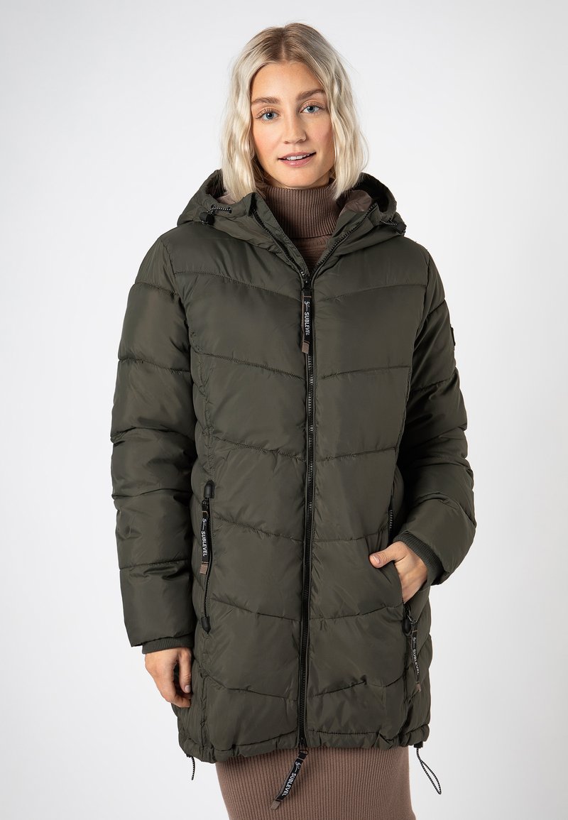 Frau in einer olivgrünen, gesteppten Winterjacke mit Kapuze, die über einem braunen, gerippten Rollkragenkleid steht und eine Hand in der Taschen der Jacke hat.