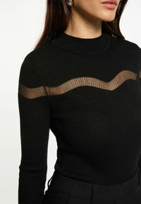 Top lungo a maniche nere con costine, caratterizzato da un pannello trasparente ondulato sulla parte superiore del petto, che riflette una texture luminosa. Design aderente, scollatura a girocollo.