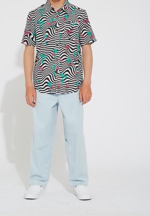 Kurzarmhemd mit schwarz-weißen wellenförmigen Streifen, verziert mit rosa Flamingos und grünen Palmen. Hellblaue Jeans und weiße Turnschuhe.