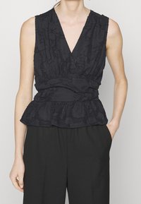 Musta ilmanhiha toppi, jossa on syvä V-pääntie, teksturoitu kukkakuosi, kerrostettu vyötärö ja peplum-helma, yhdistettynä mustiin löysiin housuihin.