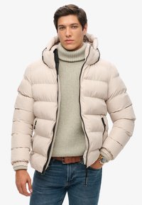 Chaqueta acolchada de color beige claro con capucha, que presenta secciones acolchadas horizontales, cremalleras negras y bolsillos laterales, llevada sobre un suéter gris.