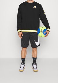 Svart sweatshirt med gula detaljer på ärmarna, gråa shorts med en vit Nike-logga och svarta sneakers. Håller en blå och grön basketboll.
