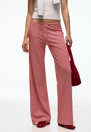 Kvinde iført røde og hvide smalle stribede bukser med vide ben, hvid cropped top, holder en rød taske og har røde sko på.