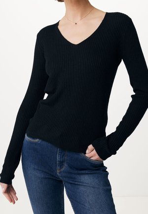 Femme portant un pull noir à manches longues à côtes avec un col en V et un jean bleu, une main dans la poche, sur fond blanc.