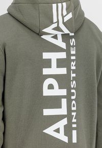 Sudadera con capucha verde oliva con texto vertical blanco "ALPHA INDUSTRIES" impreso a lo largo del lado; tejido suave y texturizado.