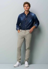 Dunkelblauses Denim-Hemd mit Knöpfen und aufgerollten Ärmeln, kombiniert mit hellbeige Chinos und weißen Turnschuhen, vor einem neutralen Hintergrund.