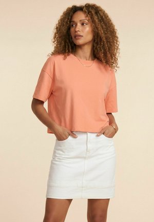 Femme aux cheveux bouclés porte un top à manches courtes couleur pêche et une jupe blanche, debout les mains dans les poches de la jupe devant un fond uni.