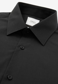 Camisa negra de botones con cuello puntiagudo, que presenta una textura suave y botones de plástico negro. La etiqueta dice "NEXT TAILORING."