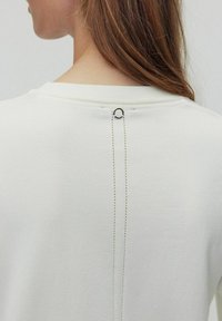 Hvid sweatshirt med rund halsudskæring og en dekorativ bagsøm med et metalringdetalje. Blød tekstur og minimalistisk design.