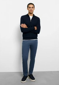 Marineblauer Strickpullover mit Reißverschluss, hellblaue Slim-Fit-Hose und schwarze Turnschuhe. Glatte Textur, klassisches Design mit Rundhalsausschnitt und Reißverschluss-Detail.