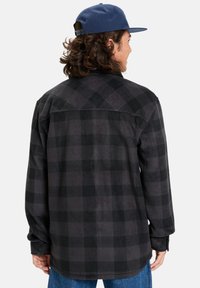 Camisa a cuadros en negro y gris para hombre, hecha de tela suave, con mangas largas, cierre de botones en la parte delantera y un dobladillo ligeramente curvado.