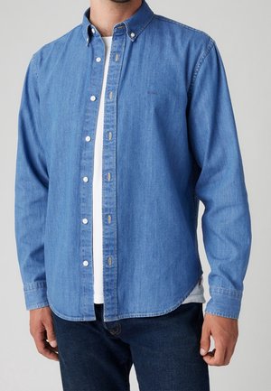 Homme portant une chemise en jean bleue à boutons sur un t-shirt blanc avec un jean bleu foncé sur un fond uni.