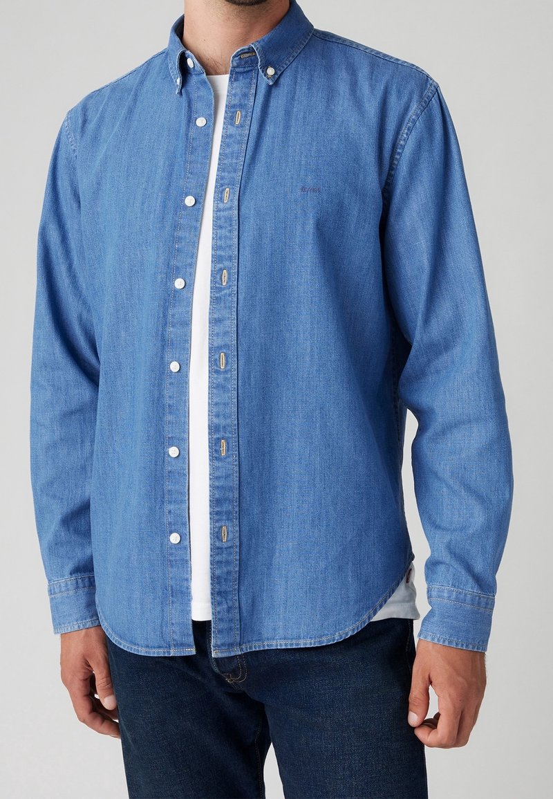 Homme portant une chemise en jean bleue à boutons sur un t-shirt blanc avec un jean bleu foncé sur un fond uni.