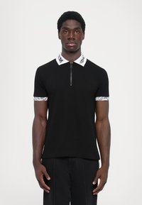 Svart polo-skjorte med glidelås på kragen, hvit logo på kragen og mansjettene, og tettsittende design. Glatt stoff med korte ermer.