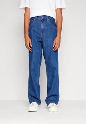 Persoon die losse blauwe spijkerbroek, witte sneakers en een wit T-shirt met korte mouwen draagt, staand tegen een effen lichte achtergrond.