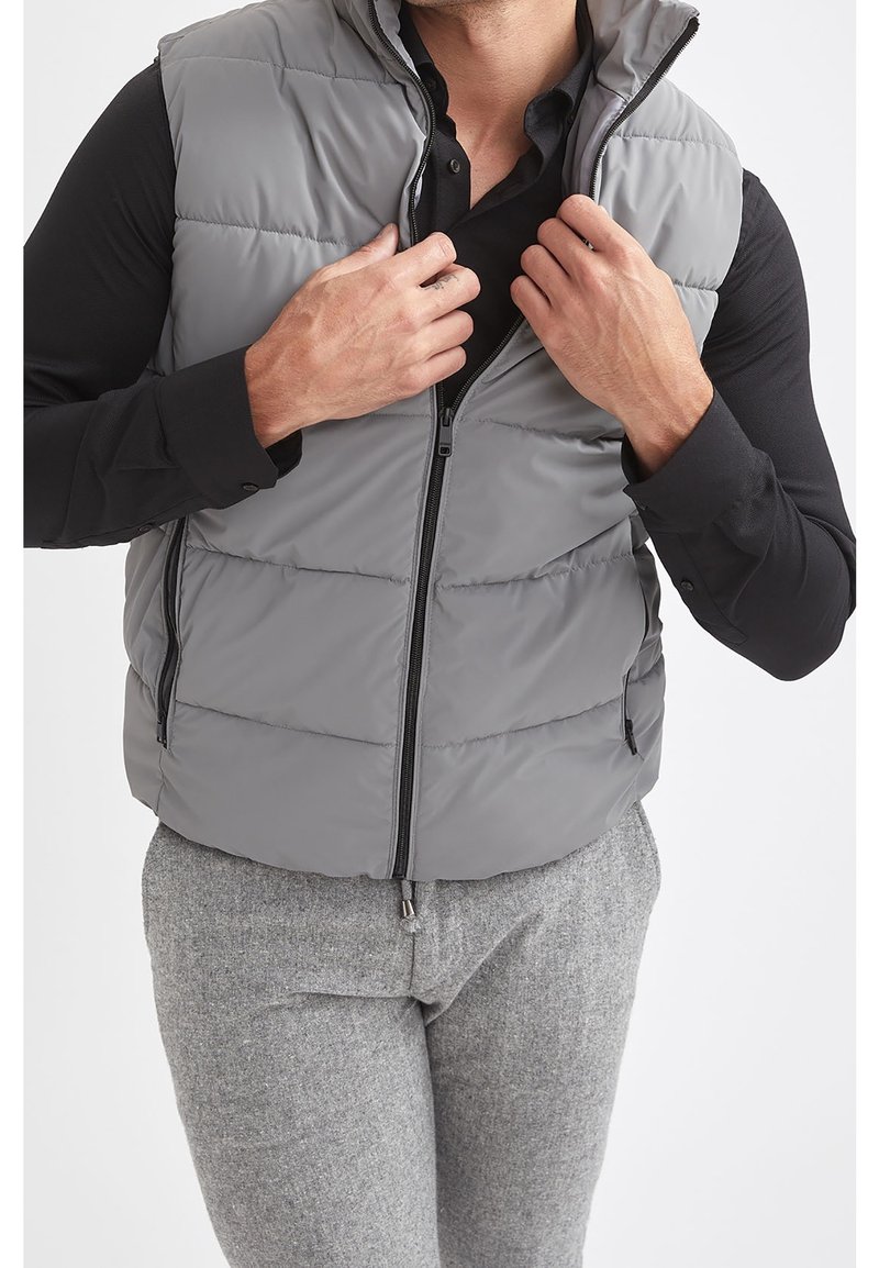 Homme portant un gilet matelassé gris sur une chemise noire et un pantalon gris, fermant la fermeture éclair du gilet avec les deux mains.