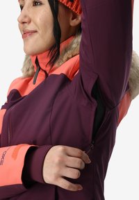 Giacca viola e arancione con colletto in pelliccia sintetica, ventilazione con cerniera sotto le ascelle e tessuto a contrasto sulle maniche. Cuciture dettagliate visibili.