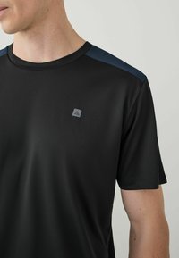 Sort atletisk t-shirt med korte ærmer og rund halsudskæring. Har marineblå detaljer på skuldrene og et lille logo på brystet.