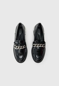 Μαύρα μοκασίνια με υφή κροκόδειλου, ασημένια αλυσίδα διακοσμητικό στο λουράκι, στρογγυλεμένη μύτη και λαστιχένια σόλα. Ορατή η εσωτερική επένδυση με λείο ύφασμα.