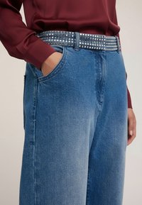 Ręka w kieszeni, ubrana w niebieskie dżinsy z nitowanym czarno-białym paskiem, zestawiona z bordową koszulą z długim rękawem.